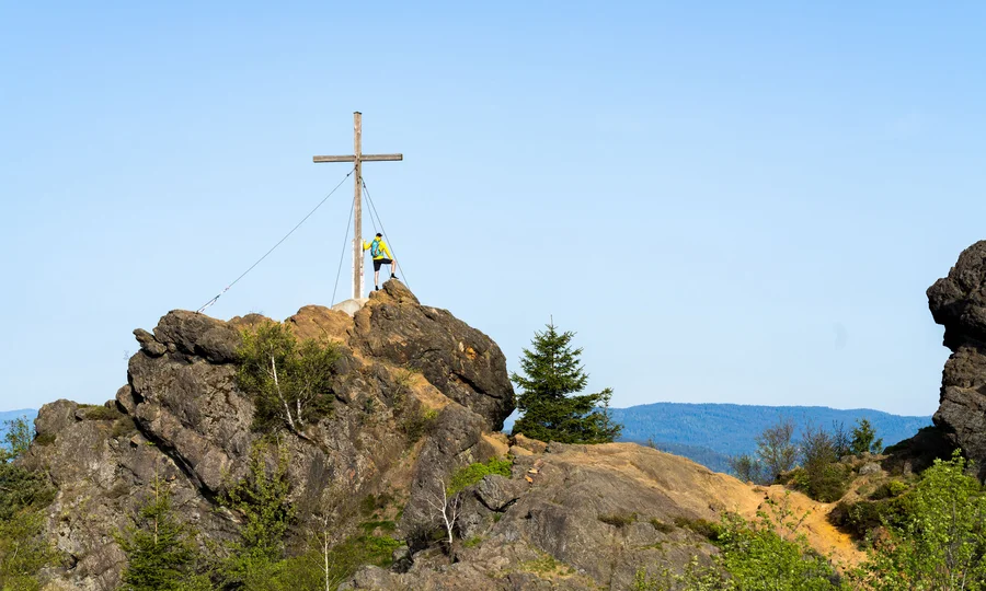 Wanderer steht an einem Gipfelkreuz auf felsigem Berg bei klarem Himmel