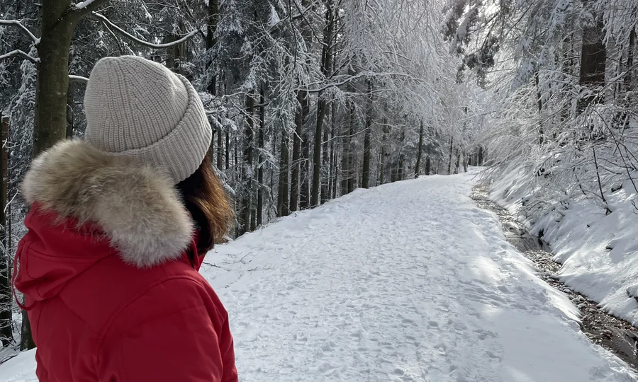 Frau in rotem Mantel und Mütze wandert auf verschneitem Waldweg