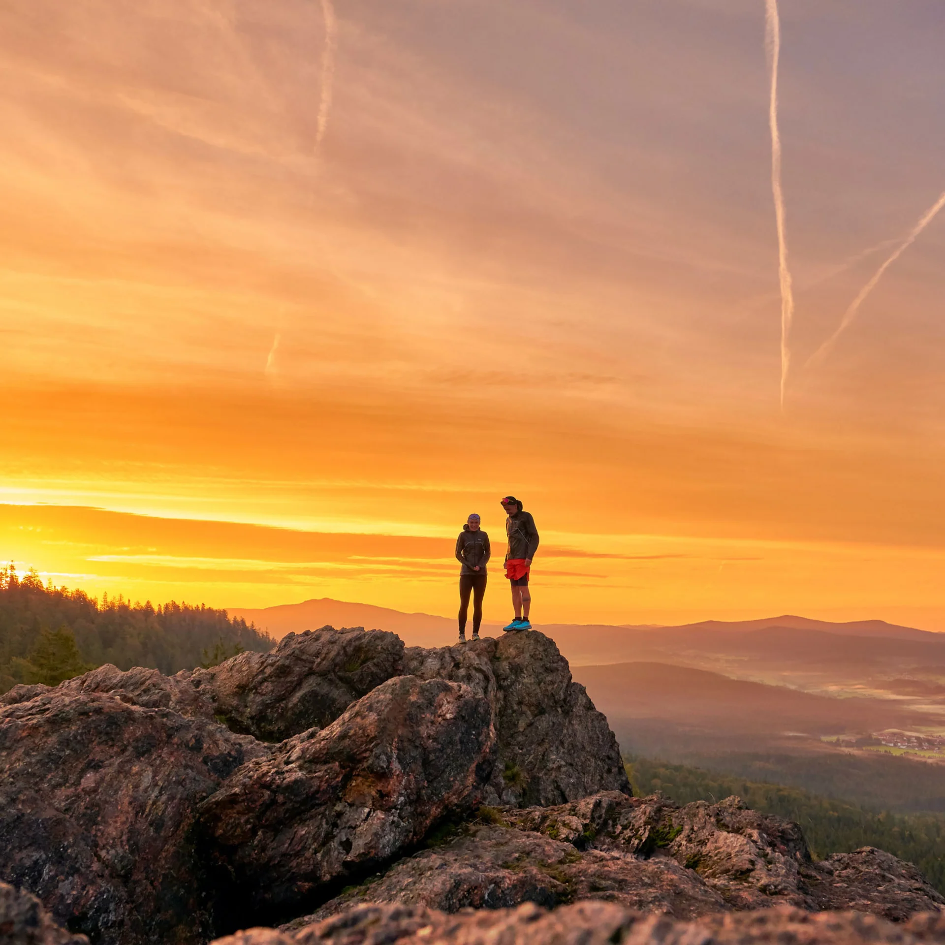 Zwei Wanderer stehen auf Felsen bei Sonnenuntergang mit Blick auf Tal und Berge