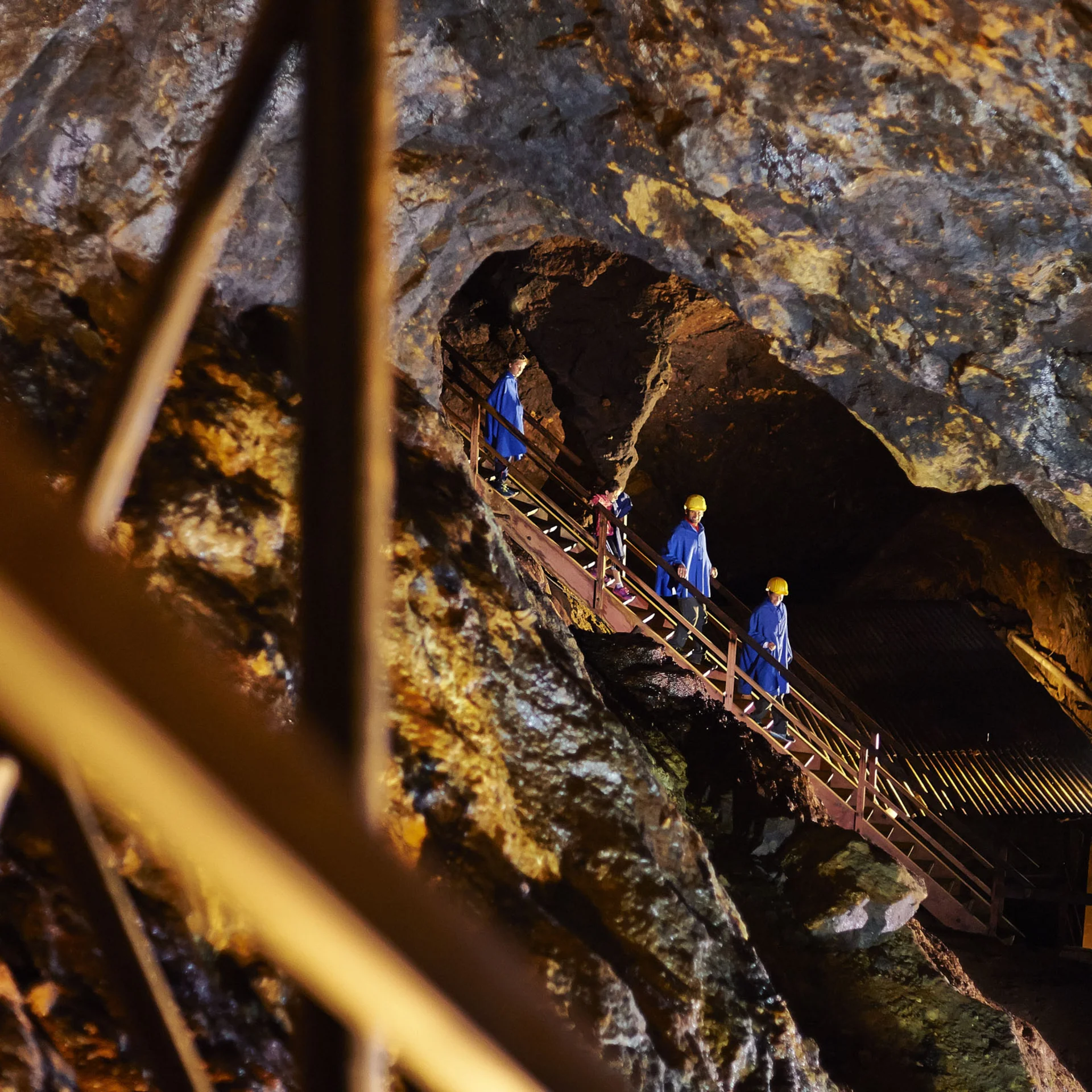 Besucher in blauen Anzügen und Helmen steigen eine Treppe in einer beleuchteten Höhle hinab