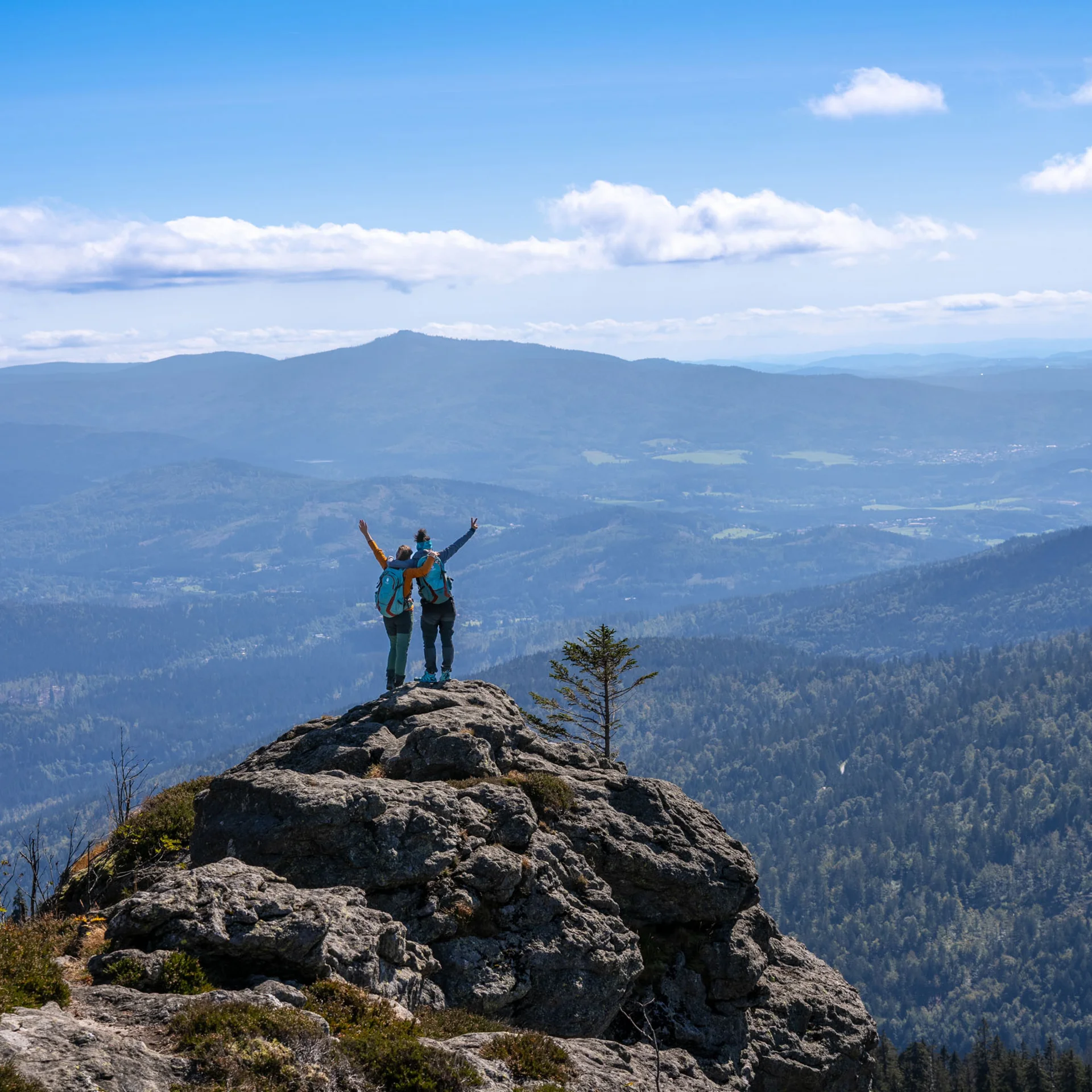 Zwei Wanderer auf einem Felsen mit Blick auf bewaldete Berge und blauen Himmel