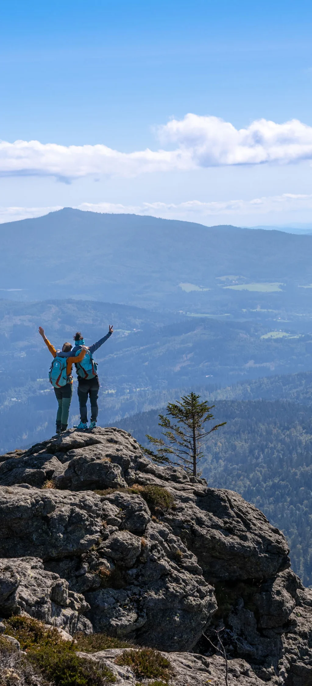 Zwei Wanderer auf einem Felsen mit Blick auf bewaldete Berge und blauen Himmel