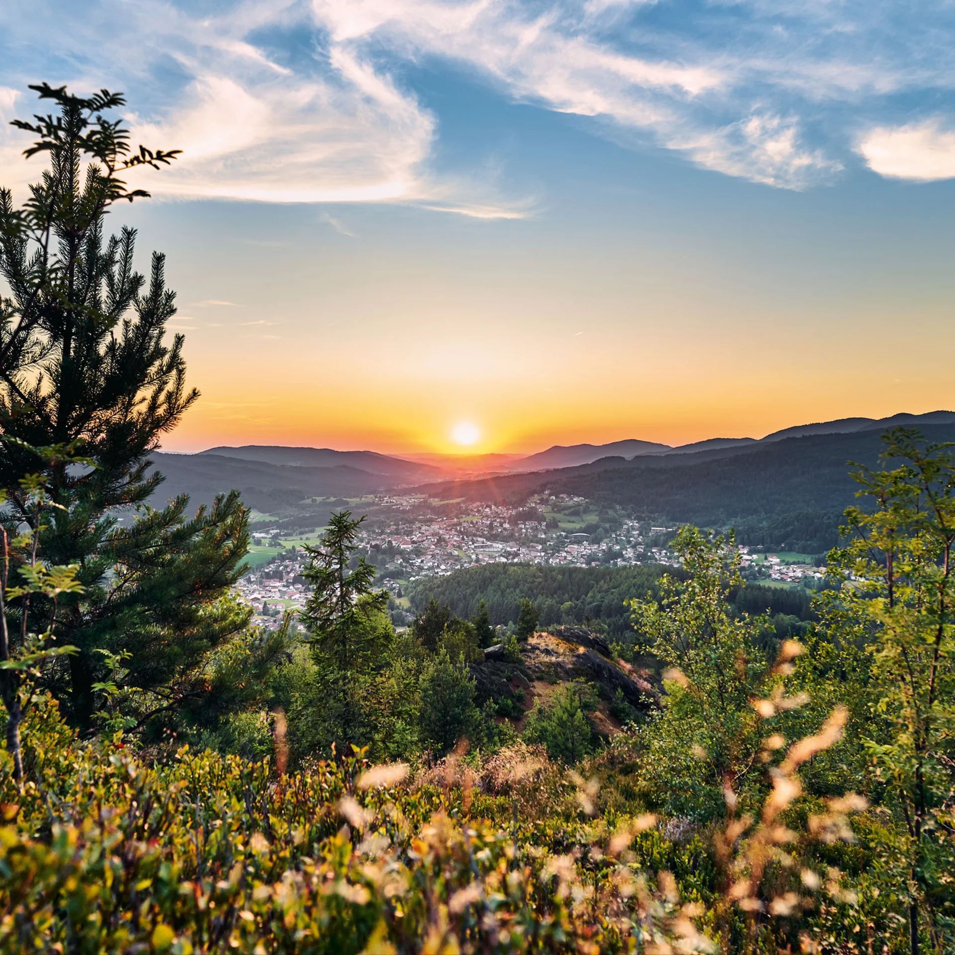 Sonnenuntergang über bewaldeten Hügeln mit Blick auf ein Tal und eine Stadt