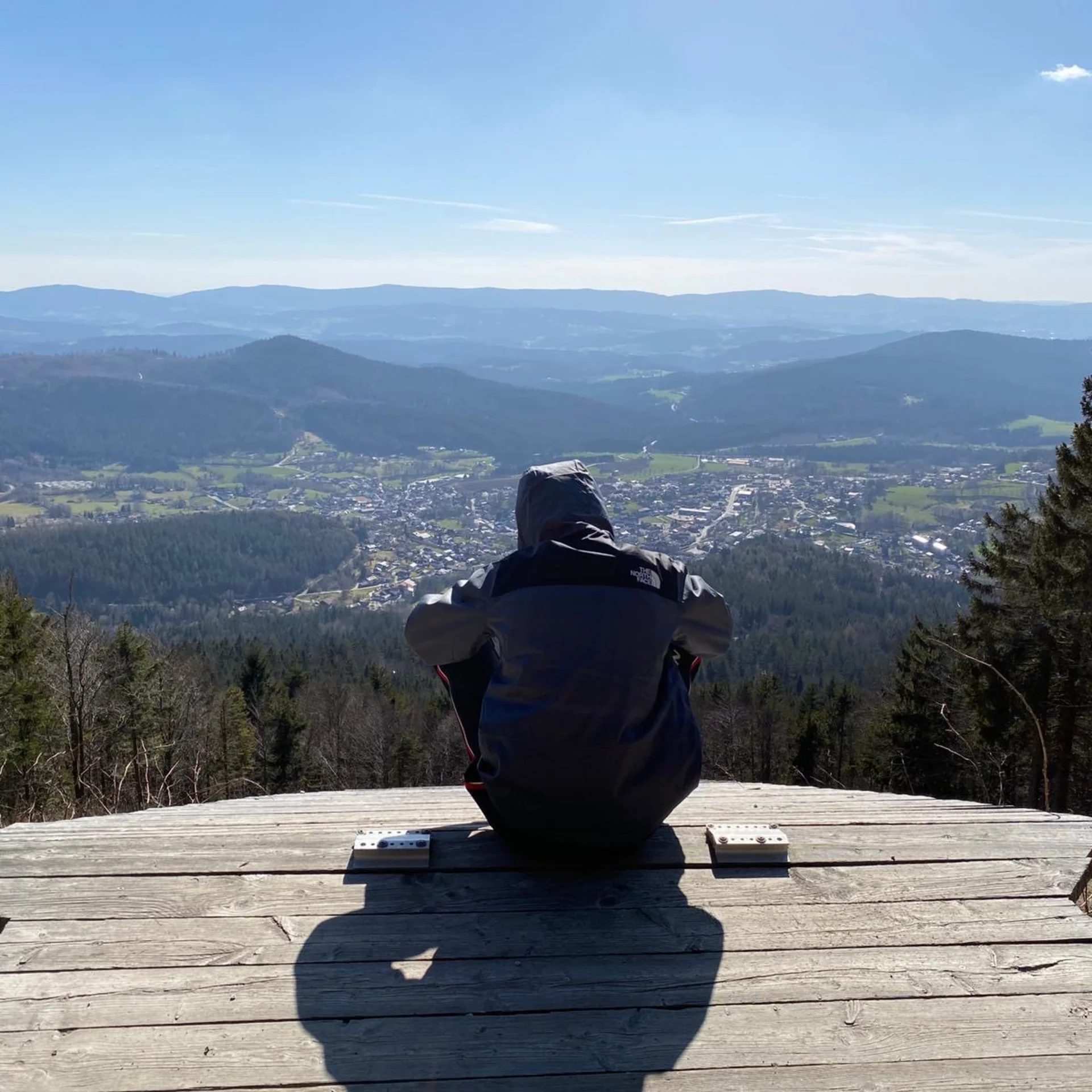 Person sitzt auf Holzplattform und blickt auf Tal und Berge bei klarem Himmel
