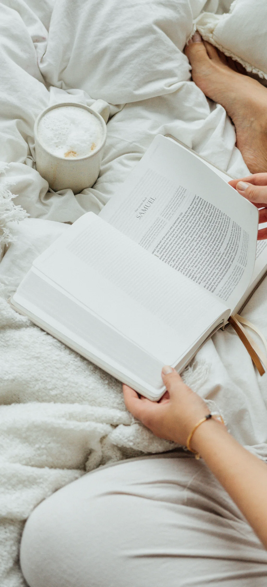 Person liest Buch im Bett mit Kaffee auf weißer Decke