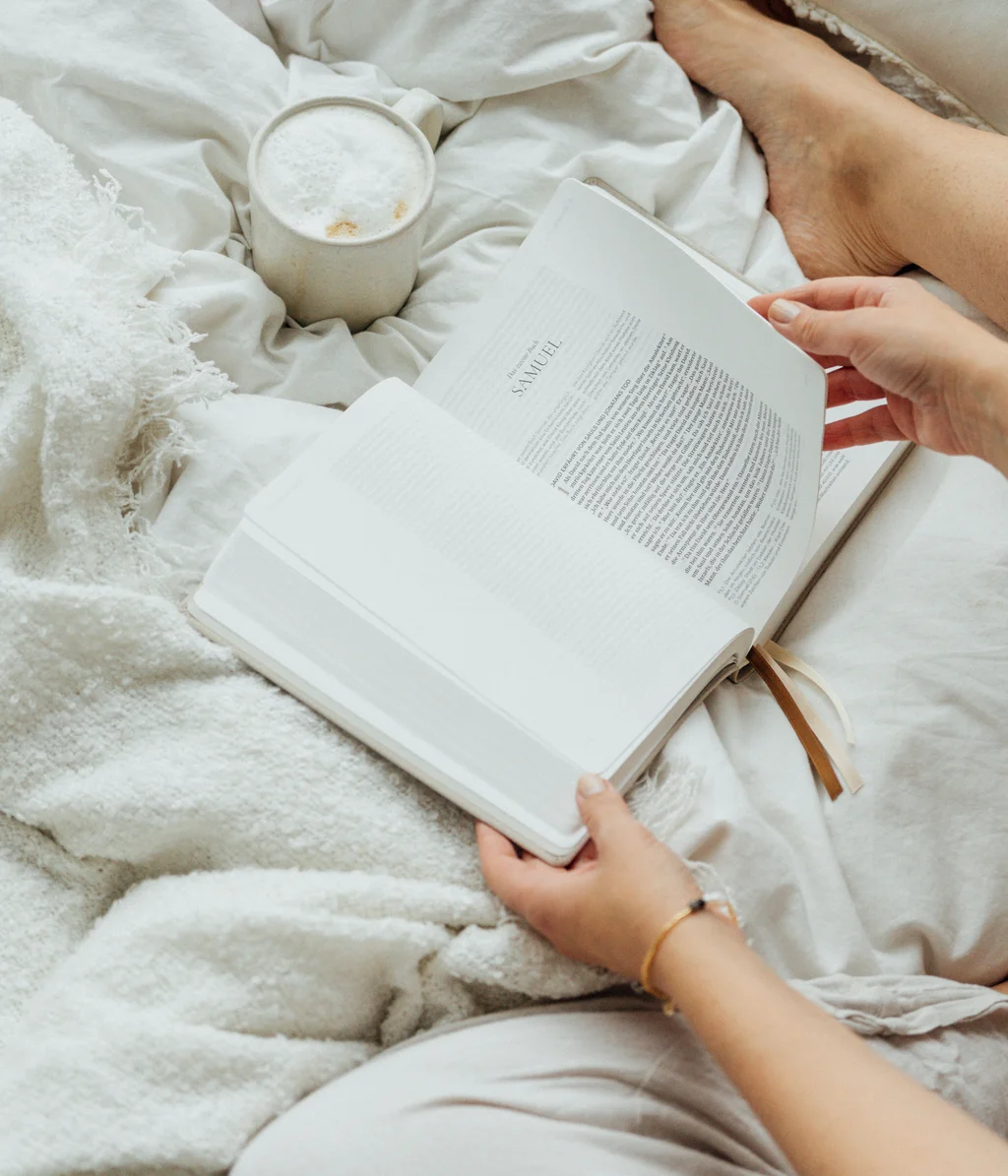 Person liest Buch im Bett mit Kaffee auf weißer Decke