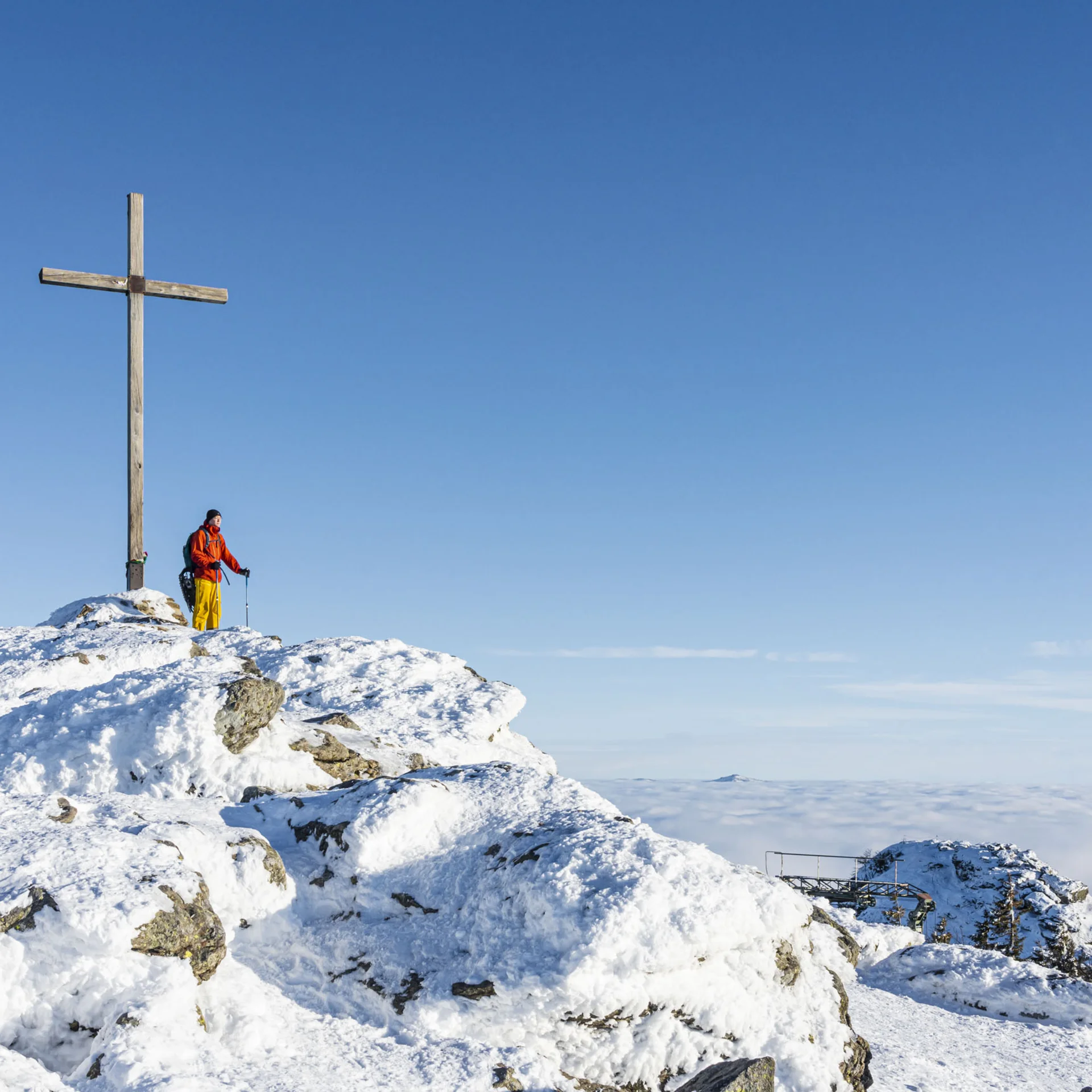 Wanderer in bunter Kleidung auf schneebedecktem Gipfel neben großem Holzkreuz